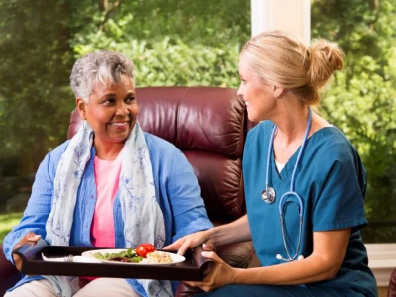 Caregiver serving a meal to a smiling senior woman at home, representing compassionate senior home care in North Las Vegas Nevada